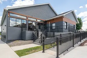 Image of a modern house with gray exterior walls and black railings, featuring a glass door and windows, under a blue sky with some clouds, with a walkway and stairs leading up to the entrance