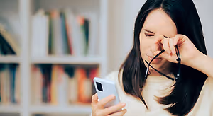 A woman holds a phone and her glasses while closing her eyes with her hand, likely due to eye strain.