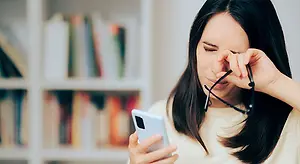 A woman holds a phone and her glasses while closing her eyes with her hand, likely due to eye strain.