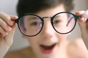A young girl is smiling and holding up a pair of black glasses with clear lenses, showing her reflection and the glasses' frame.