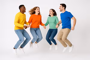 Four people wearing yellow, orange, green, and blue shirts are jumping and holding hands in the air. The two women are wearing jeans and the two men are wearing khaki pants. The background is white.