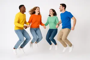 Four people wearing yellow, orange, green, and blue shirts are jumping and holding hands in the air. The two women are wearing jeans and the two men are wearing khaki pants. The background is white.