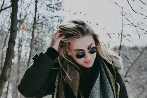 Blonde woman wearing sunglasses and a fur collared jacket standing in a forest.