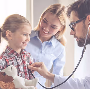 A doctor is examining a child with a stethoscope while the child's mother looks on, all three smiling at the camera.