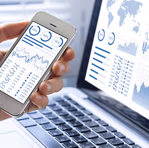 A person is holding a mobile phone with graphs and charts on the screen, and it is placed on top of a laptop with a keyboard and a monitor displaying a world map and financial data.