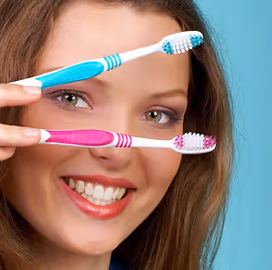 A woman holding two toothbrushes, one with pink and white bristles and the other with blue and white bristles, is smiling and looking at the camera.