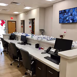 An office space has a long desk with computer monitors, keyboards, and telephones. A large monitor is mounted on the wall, and there are vases with flowers and balloons on the desk. There are also two doors with signs and a camera on the ceiling.