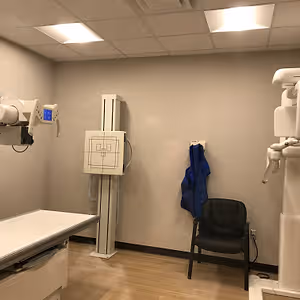 A room with a medical X-ray machine, a chair, and a blue jacket hanging on the wall.