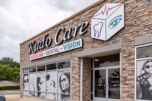 The outside of the Kud Care building with a glass door, a logo, and a sign with text and images of people and sunglasses.
