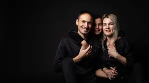 A man and two women are sitting close together, smiling and posing for a photo with a dark background. They are all dressed in black.