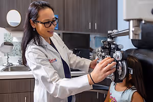 A woman doctor in a white coat examines a young girl's eyes with an ophthalmoscope in an exam room.