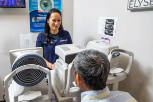 A woman is looking into a machine while a man is looking at her and another machine in an eye clinic.