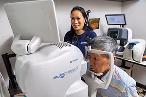 An older man is getting his eyes checked by a woman in an eye clinic with monitors and medical equipment in the background.