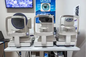 Three white phoropters, which are used in eye exams, are displayed on a table in a medical office. There is a television and a poster on the wall behind the phoropters. A laptop and a box are on the left side of the table, and a container is on the right side.