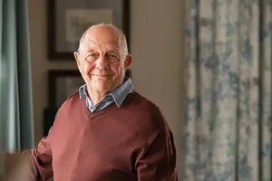 An elderly man wearing a maroon sweater stands and smiles by a window with a curtain in the background.