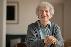 An elderly woman with white hair smiles while leaning on a wooden cane inside a house.