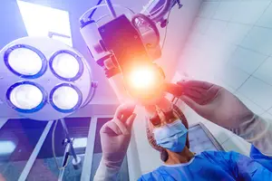 A person in a surgical mask and blue scrubs holding an ophthalmoscope with a bright light in a hospital room.