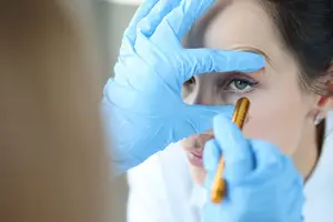 A woman in a white lab coat is examining a patient's eye with a gold-colored object.