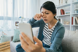 woman sitting on a couch holding a tablet and looking at the camera while rubbing her eyes