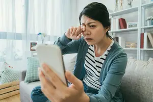 woman sitting on a couch holding a tablet and looking at the camera while rubbing her eyes