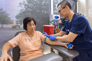 An elderly woman is sitting on a chair and having her blood drawn by a male healthcare worker in a clinic.