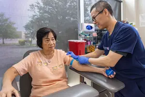 An elderly woman is sitting on a chair and having her blood drawn by a male healthcare worker in a clinic.