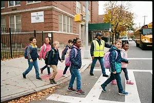 Children in school uniforms are crossing the street with a school safety officer and a school bus is in the background.