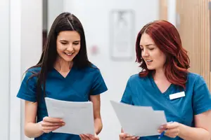 Two women in blue uniforms are looking at papers and smiling in a hospital hallway.
