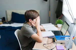 A boy is sitting on a chair in a room and seems to be thinking while keeping his hands on his cheeks.