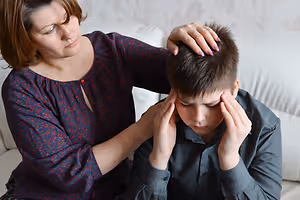 A woman sitting on a couch is comforting a young boy who is experiencing a headache, she has her hand on his head while he has his eyes closed and is holding his head in pain.