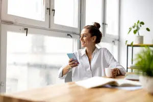 Woman sitting at a desk with a cell phone and coffee cup in her hands, smiling.