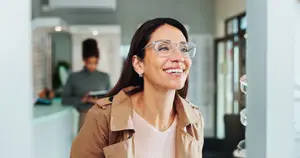 A smiling woman in glasses is standing in front of a white wall in a shop, with a blurred image of a woman standing in front of a desk in the background.