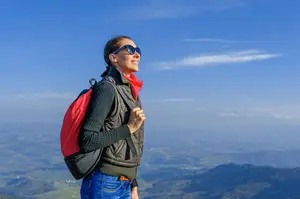 A woman wearing a backpack and a red and black jacket is standing on a mountain peak, smiling and looking at the view.