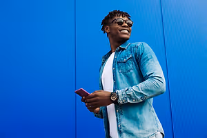A smiling man wearing glasses and a denim jacket stands in front of a blue wall, holding a pink cell phone. He seems to be taking a selfie. 