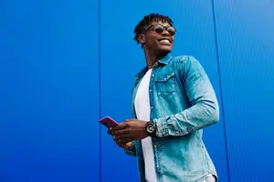 A smiling man wearing glasses and a denim jacket stands in front of a blue wall, holding a pink cell phone. He seems to be taking a selfie. 