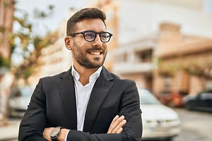 A smiling man in a suit and glasses stands on a street with his arms crossed.