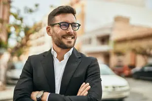 A smiling man in a suit and glasses stands on a street with his arms crossed.