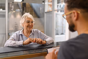 A smiling elderly woman in glasses is talking to a man in an office setting
