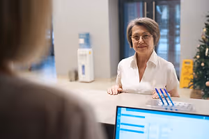 An older woman wearing glasses is smiling and looking at something on the right. She is probably inside an office. In front of her is a desk with a monitor, a bottle, and a pen. Behind her is a blurry person, a Christmas tree, a water dispenser, and a glass door. On the right is a yellow sign on the wall.