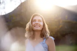 A woman with blonde hair is smiling in front of a mountain and trees with a bright sunlight shining on her face