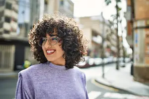Woman with curly hair, wearing glasses, smiling and standing in a street
