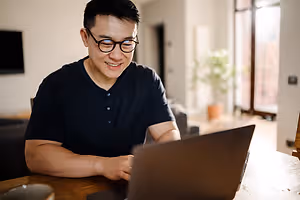 A smiling man is sitting on a chair, using a laptop in a room