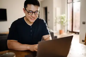 A smiling man is sitting on a chair, using a laptop in a room