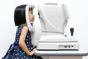 A young girl in a blue polka dot dress is sitting and looking into an eye examination machine.