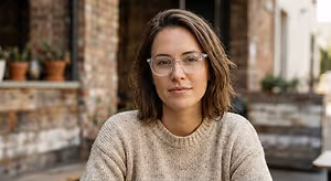 A woman wearing glasses and a beige sweater sits in front of a building, looking to the side.