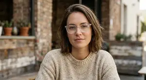 A woman wearing glasses and a beige sweater sits in front of a building, looking to the side.