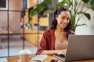 A smiling woman is sitting on a chair in front of a table with a laptop, cup, book, and phone.
