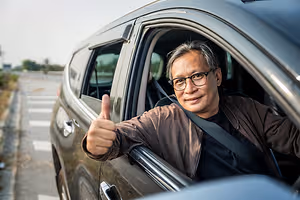 A man in a brown jacket sits in a car and gives a thumbs up while smiling.
