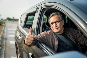 A man in a brown jacket sits in a car and gives a thumbs up while smiling.