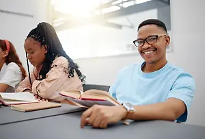 A young adult male sitting in a classroom wearing glasses and a blue t-shirt reading a book with his friends sitting behind him in the classroom.
